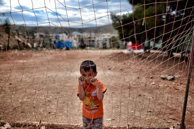 Child in orange shirt stands behind a net in an open, barren area, with tents and trees in the background.