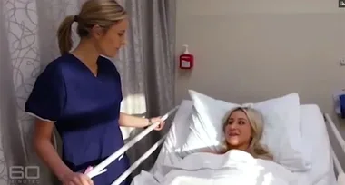 A nurse in blue scrubs stands beside a blonde woman lying in a hospital bed, conversing and smiling.