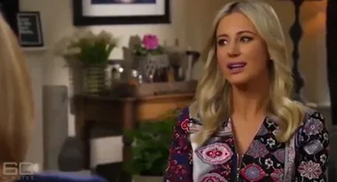 A woman with long blonde hair in a colorful patterned top speaks in a room with flowers and a framed picture.