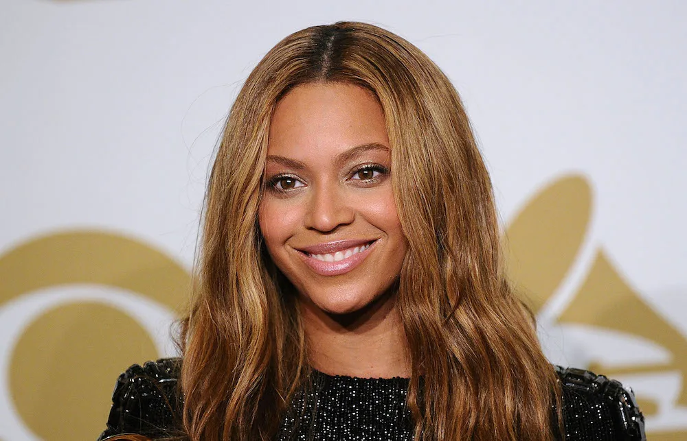 A person with long, wavy hair smiles at an event with a gold logo in the background.