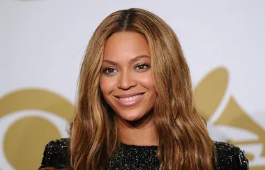 A person with long, wavy hair smiles at an event with a gold logo in the background.