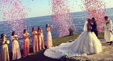 Bride and groom kiss at an outdoor beach wedding surrounded by bridesmaids and petals.