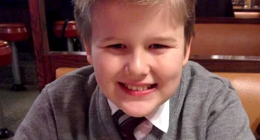 A smiling child in a gray sweater and tie sits at a booth in a diner.
