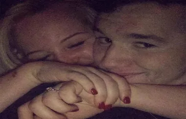 Two people cuddling closely, smiling with their faces touching and holding hands in a dimly lit setting.