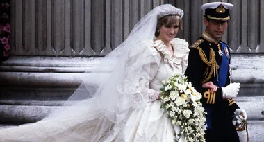 Wedding couple in formal attire, bride in a white gown with flowing veil and groom in a naval uniform, holding hands.