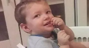 A toddler smiling, sitting in a high chair, with short brown hair, wearing a light blue shirt, indoors.