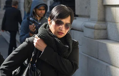 A woman in sunglasses and a black scarf walking outside, with a stone building in the background.