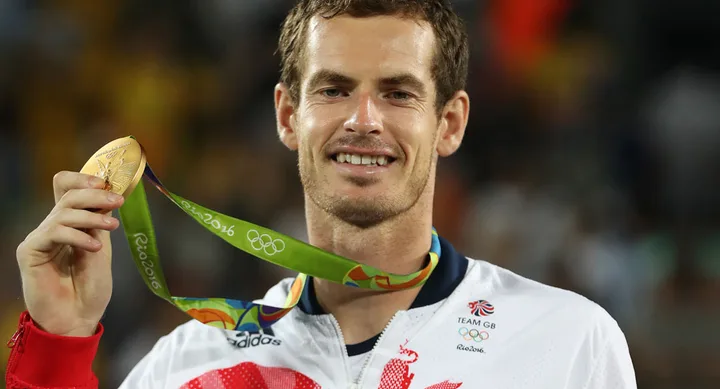 Man in athletic attire holds a Rio 2016 Olympic gold medal, smiling at the camera.