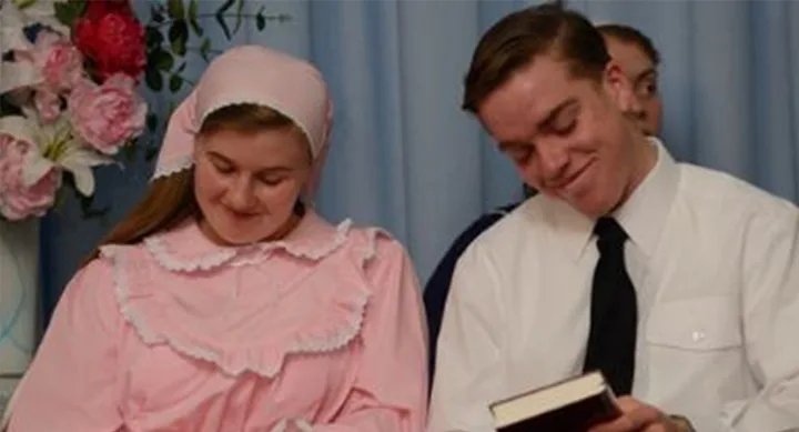 Two people sitting and smiling, one in a pink dress with a bonnet, the other in a white shirt holding a book.