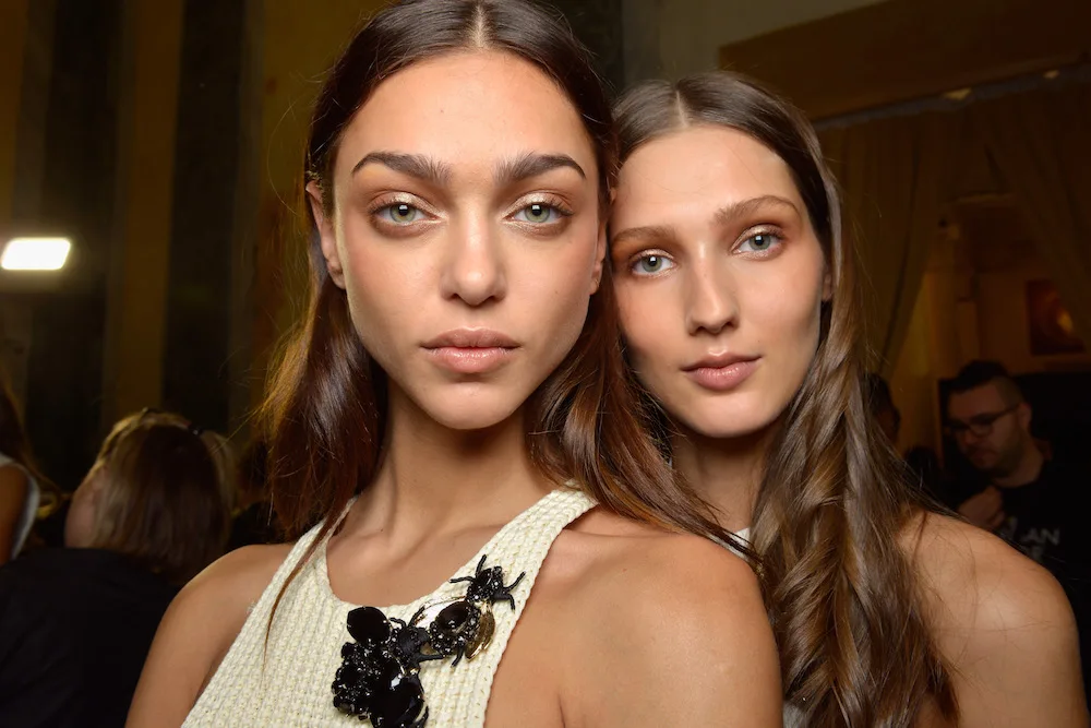 Two women with long hair posing closely in a dimly lit room, one wearing a white top with black embellishments.