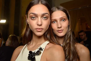 Two women with long hair posing closely in a dimly lit room, one wearing a white top with black embellishments.