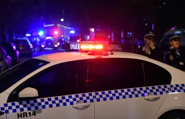 Police car with flashing lights at night, officers standing nearby on a street with emergency vehicles in the background.
