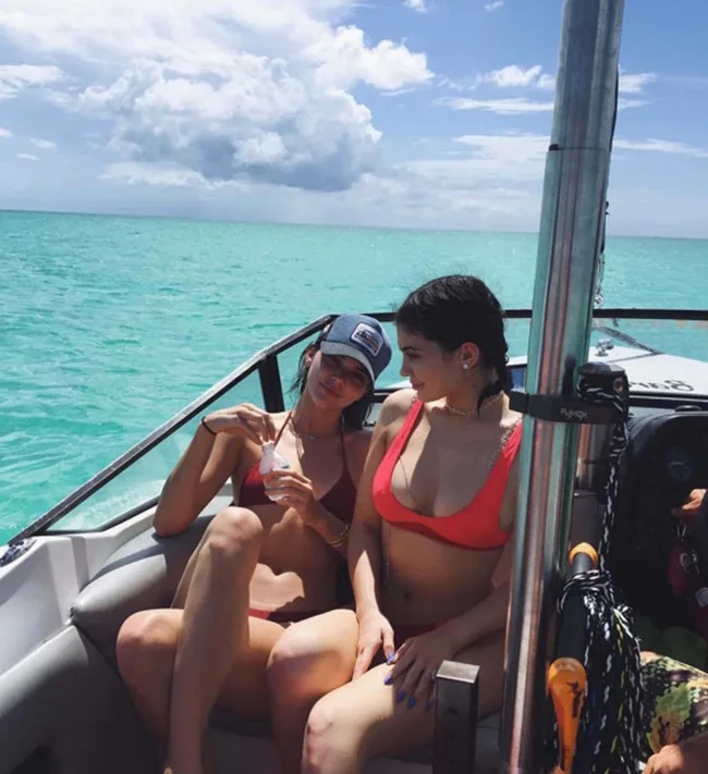 Two people in bikinis relaxing on a boat in turquoise ocean under a partly cloudy sky.