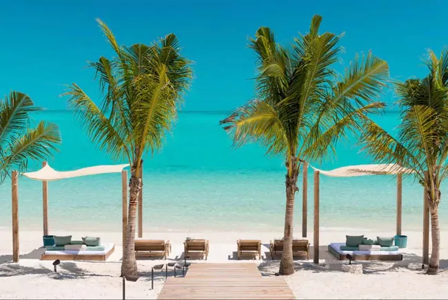 Tropical beach with palm trees, shaded lounge beds, and turquoise water under a clear blue sky.