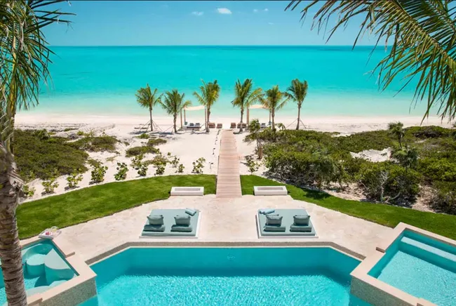 Luxury beachfront view with pool, palm trees, and turquoise ocean under a clear blue sky.