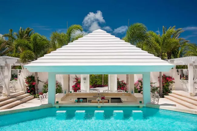 Outdoor pool bar with white pergola roof, palm trees, and vibrant flowers in the background under a clear blue sky.