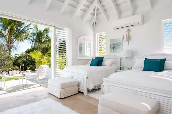 Bright bedroom with two white beds, teal pillows, and open patio doors revealing tropical garden and palm trees outside.