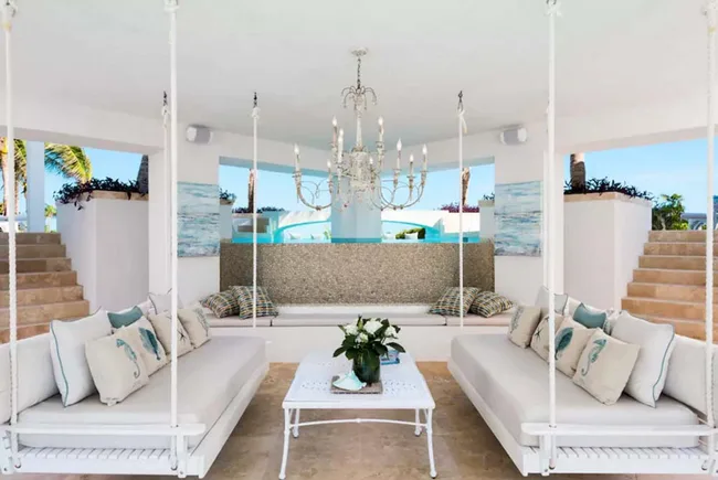 Luxurious outdoor lounge with hanging sofas, chandelier, and a central table. Bright, airy, and elegantly furnished.