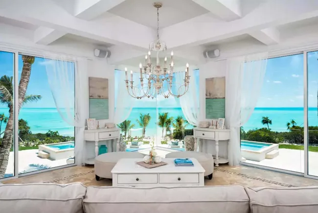 Luxurious beachside living room with ocean view, large windows, white decor, chandelier, and plush seating.