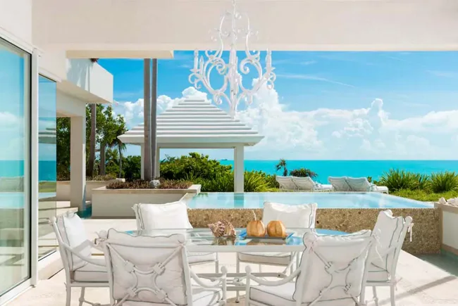 Oceanfront patio with white furniture, a glass table, pool, and ocean view under a blue sky with fluffy clouds.