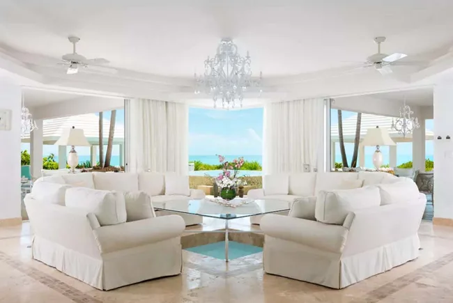 Luxurious white living room with sofas, glass table, chandelier, and ocean view through large windows.