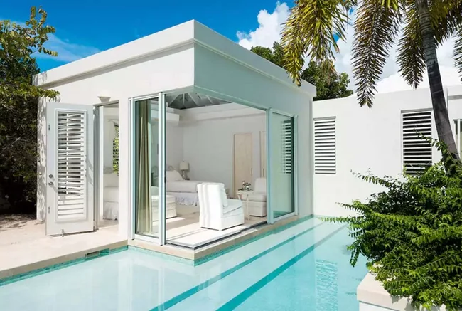 Modern white villa with large glass windows and a private pool surrounded by lush greenery under a clear blue sky.
