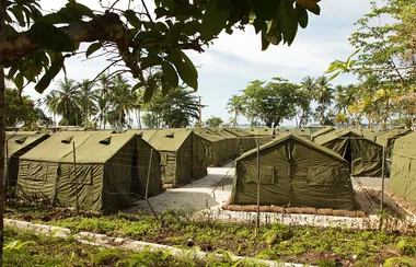 Green tents in a fenced area, surrounded by trees, with a backdrop of palm trees and ocean under a blue sky.