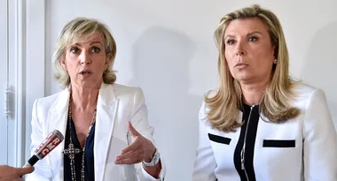 Two women in white blazers at a press event, one speaking with hand gestures, an RMC microphone visible.