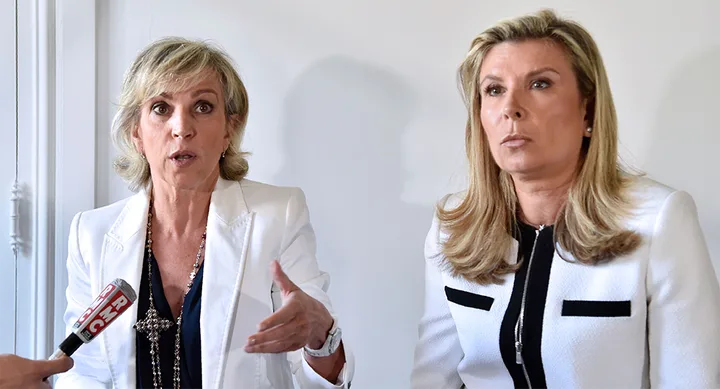 Two women in white blazers at a press event, one speaking with hand gestures, an RMC microphone visible.