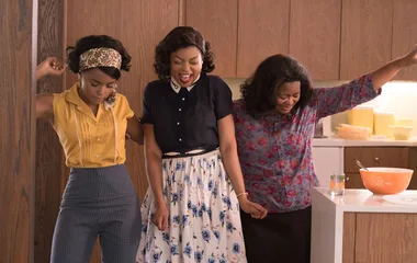 Three women in vintage clothing joyfully dance in a kitchen with orange bowl and wooden cabinets.
