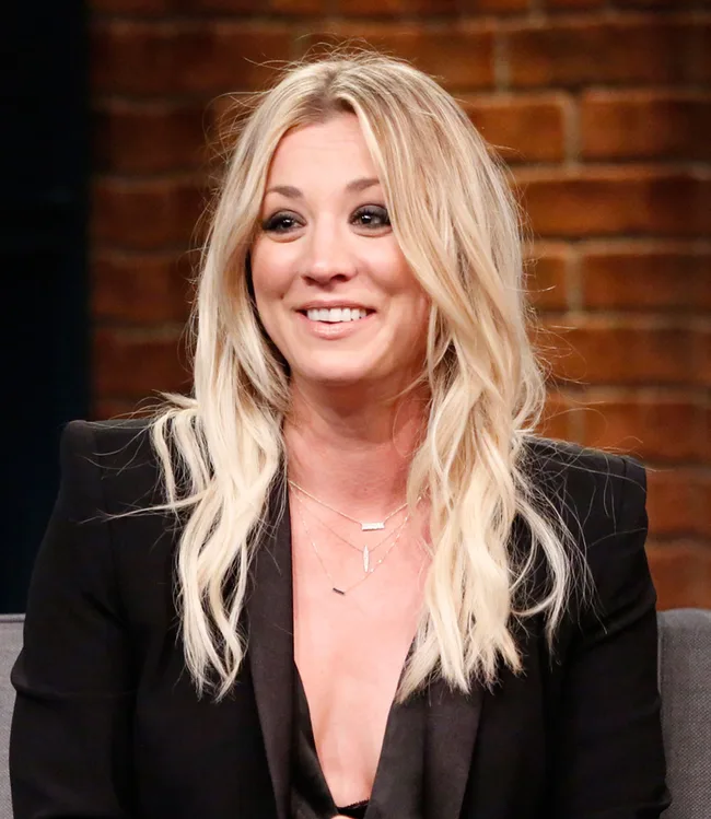 Blonde woman smiling, wearing a black outfit, sitting indoors with a brick wall backdrop.
