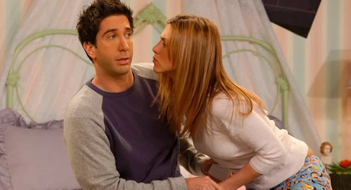 Two people sitting on a bed; one looks surprised while the other leans in closely, in a cozy bedroom setting.