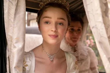 A woman in Regency-era dress with a tiara and necklace looks out of a carriage window; another woman is partially visible behind her.