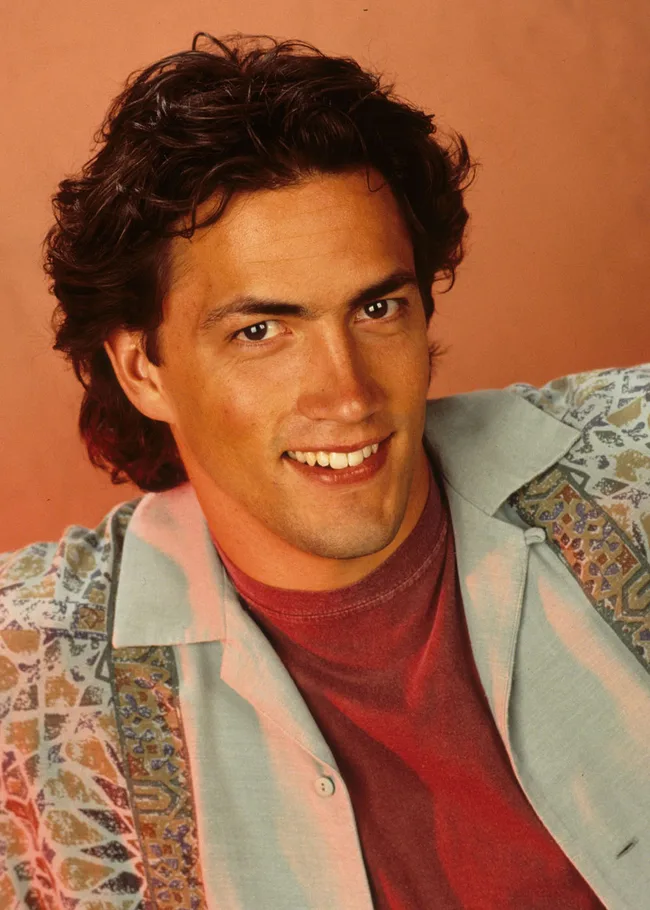 A person with dark hair smiling warmly, wearing a red shirt and patterned jacket against an orange background.