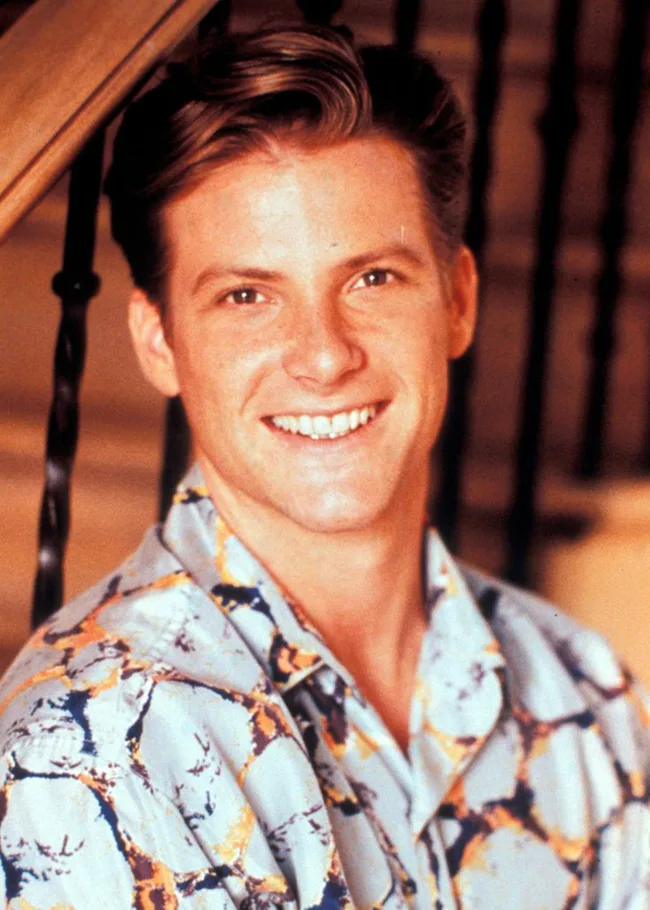 A person with short hair smiles, wearing a patterned shirt, standing indoors near a staircase.