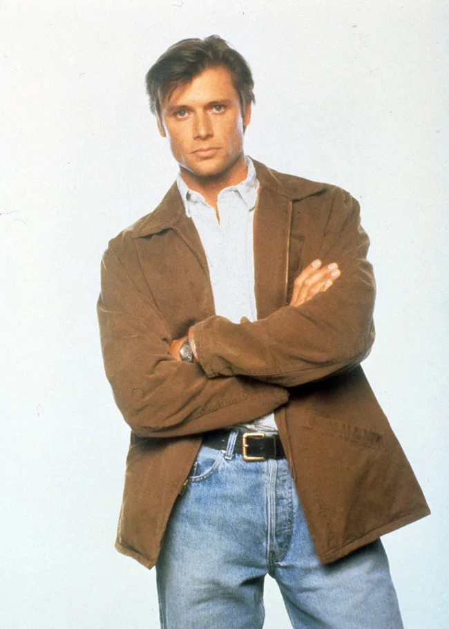 Man in a brown jacket and jeans, with arms crossed, against a light background.