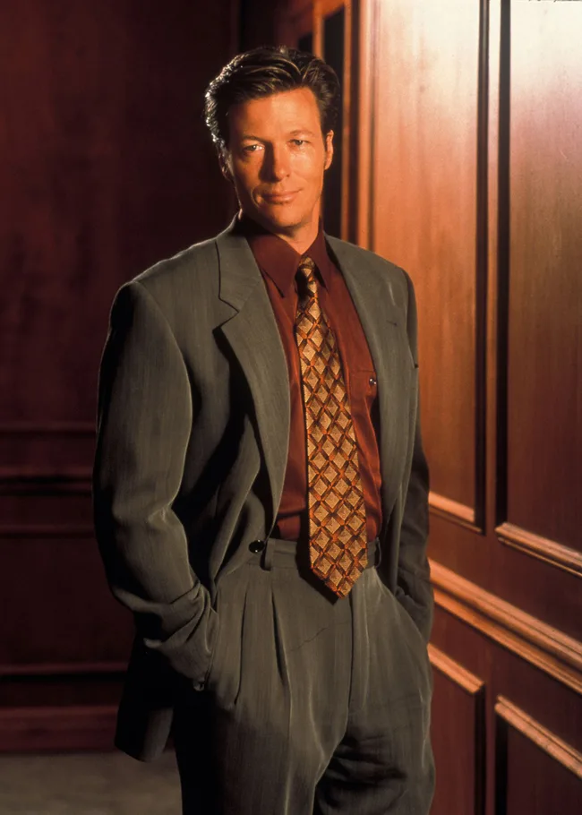 A man in a gray suit with a patterned tie stands confidently against a wooden-paneled wall.