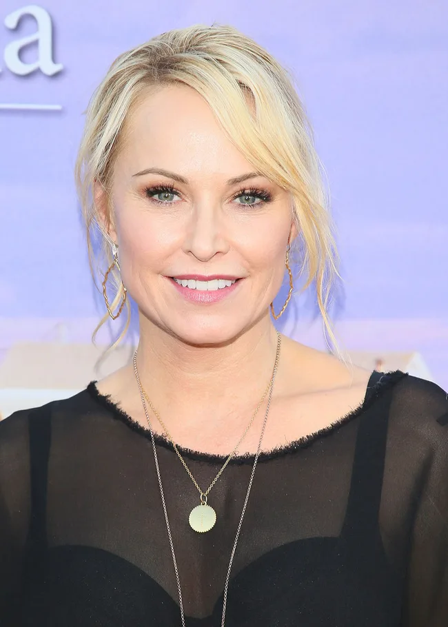 A woman with blonde hair in a black top smiles at a purple-themed event backdrop.