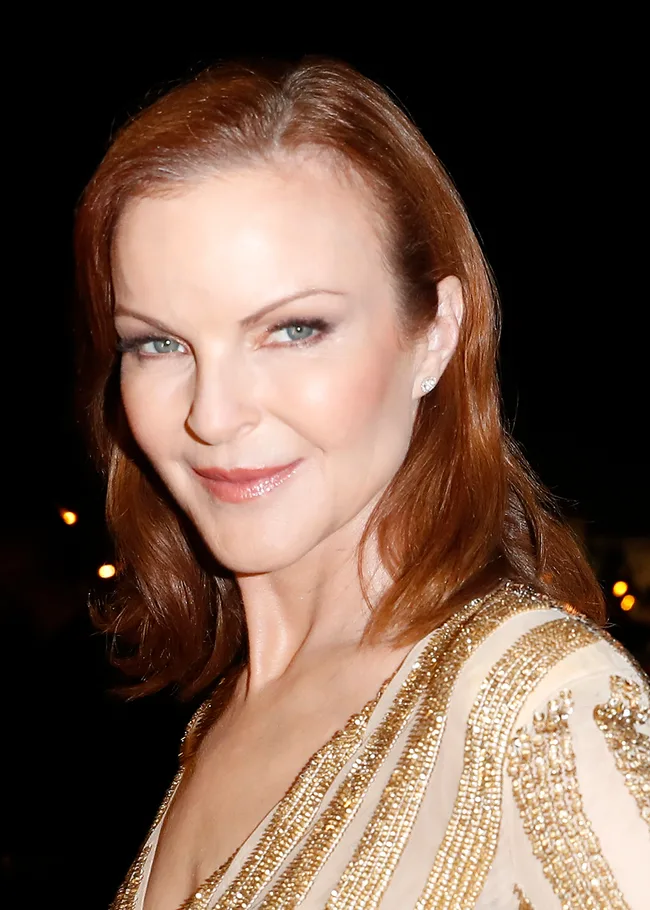 A woman with red hair smiles, wearing a gold-accented outfit, against a dark background.