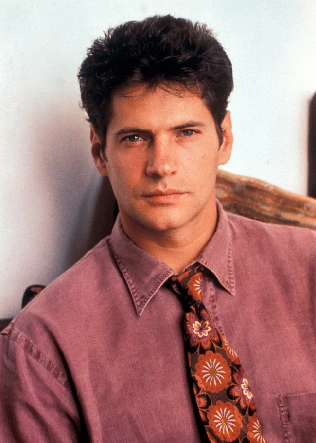 Portrait of a man with dark hair, wearing a pink shirt and patterned tie, looking at the camera.
