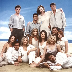 Cast of "Melrose Place" posing together on a beach, all wearing light-colored clothing against a blue sky backdrop.