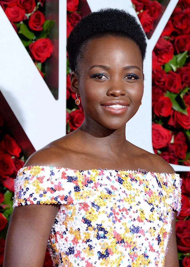 A person smiling, wearing a colorful sequined dress, with a backdrop of red roses.