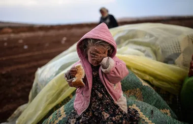 Young child in pink hooded coat covering face, holding bread, with blurred adult and large covered items in background.