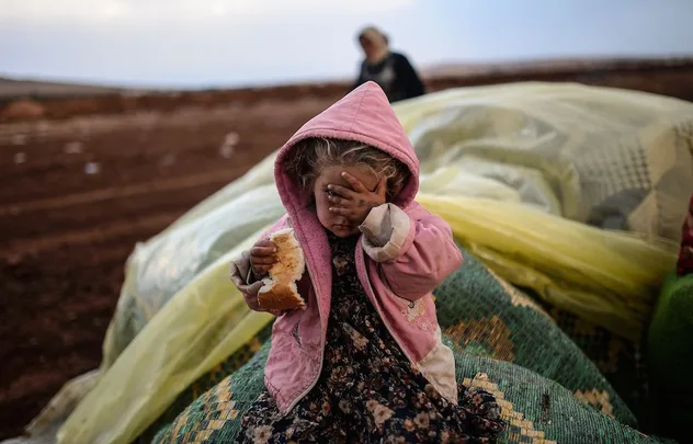 Young child in pink hooded coat covering face, holding bread, with blurred adult and large covered items in background.