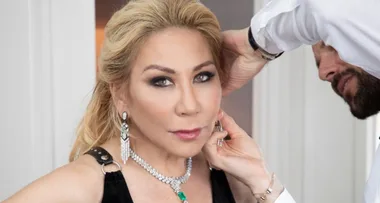 Woman getting her makeup and jewelry adjusted by a stylist, wearing a black dress and green jeweled necklace and earrings.