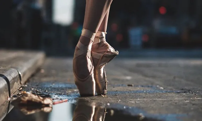 Ballerina on pointe shoes standing on a wet city street near a puddle, with blurred urban background.