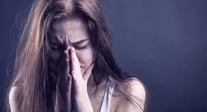A woman with long hair is looking down, appearing to be distressed, with her hands clasped near her face.