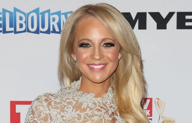 Blonde woman in a lace dress smiles at an event, standing with logos in the background.