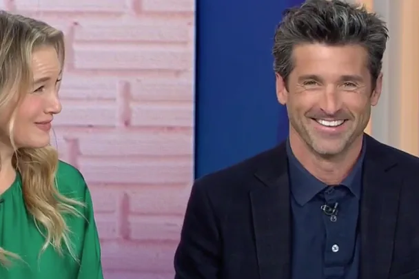 Two people smiling during a TV interview, one wearing a green top and the other in a dark blazer.