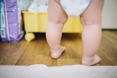 Toddler in a diaper taking steps on a wooden floor, next to a yellow cart and a pack of diapers.